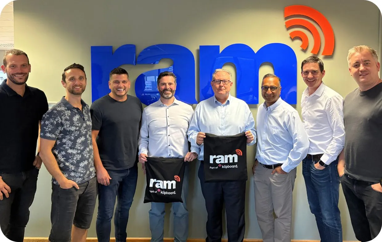 Eight men standing in front of a wall with 'ram' logo, two holding black bags with same branding.