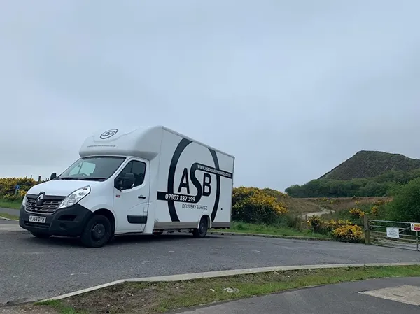 White ASB delivery van parked on road near green bushes and a small hill under a cloudy sky.