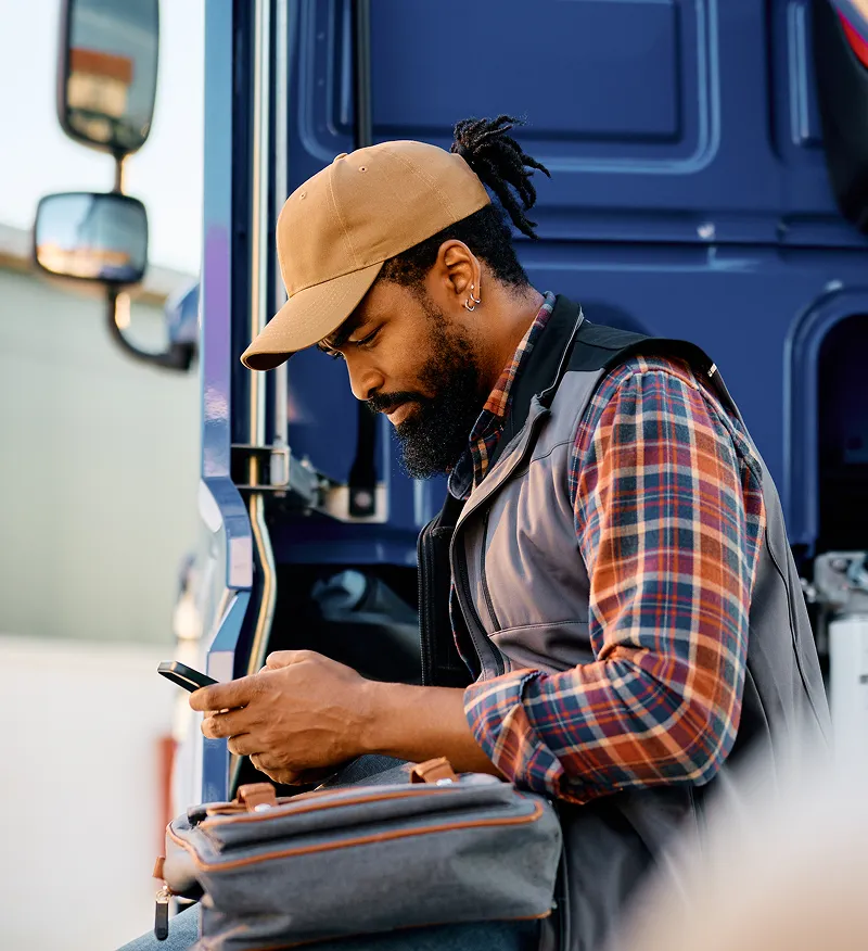 Man in a cap and check shirt sitting by a truck, looking at his smartphone with a bag on his lap.