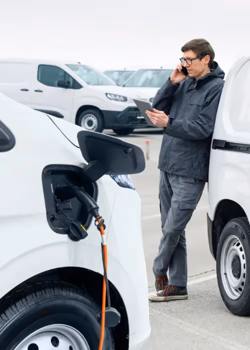 man on phone leaning on van while looking at tablet while another electric van charges