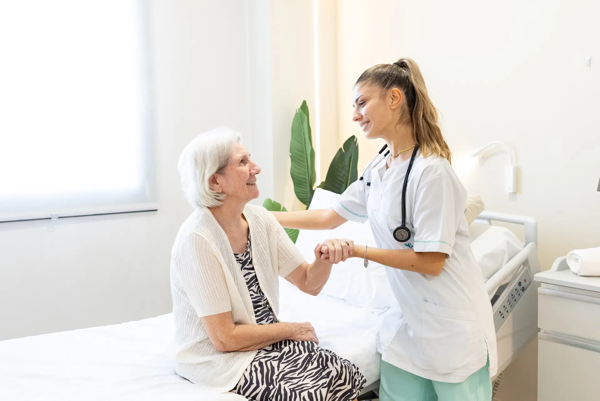 Nurse holding hands with a smiling older woman sitting on her bed