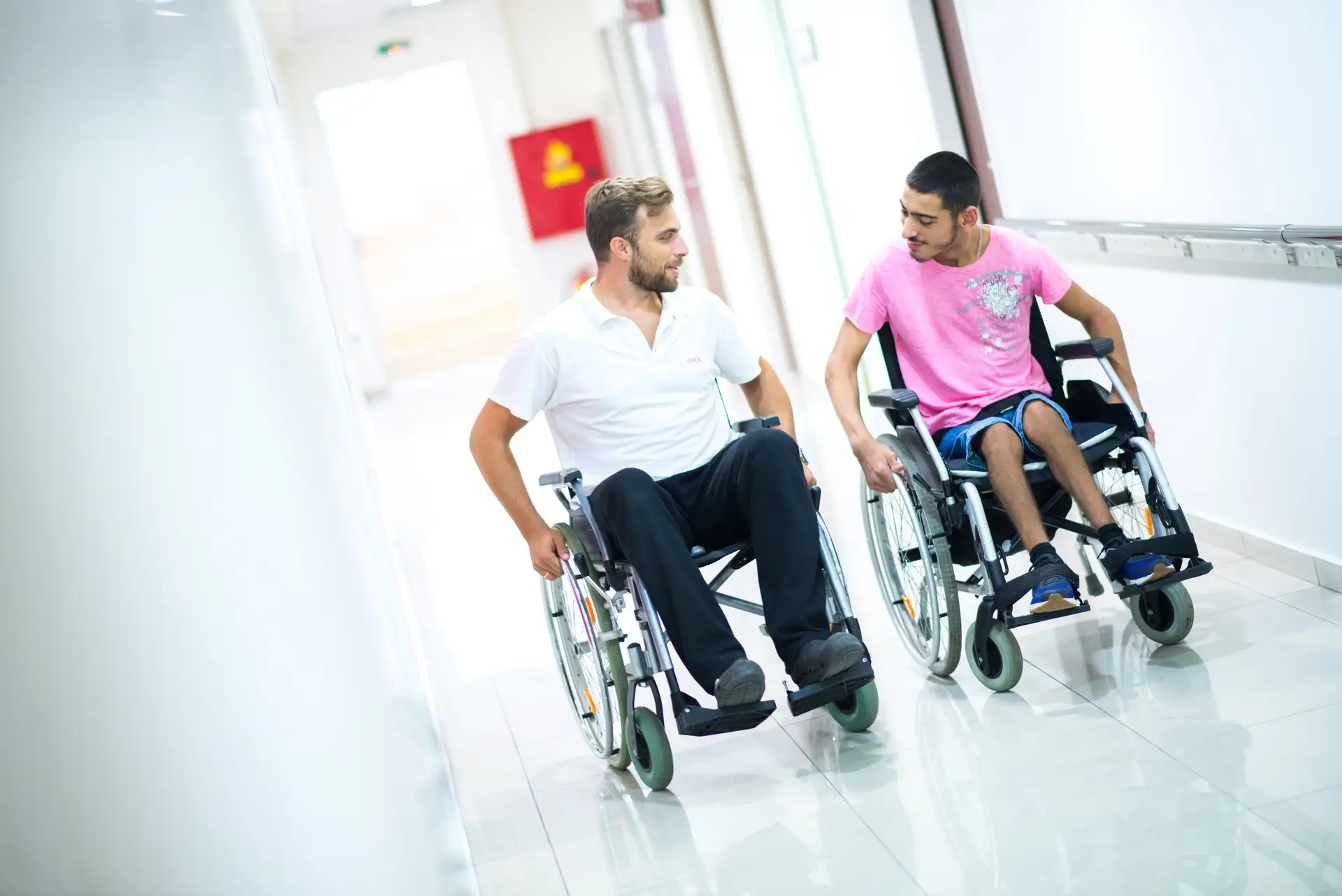 Two men in wheelchairs moving together along a bright corridor