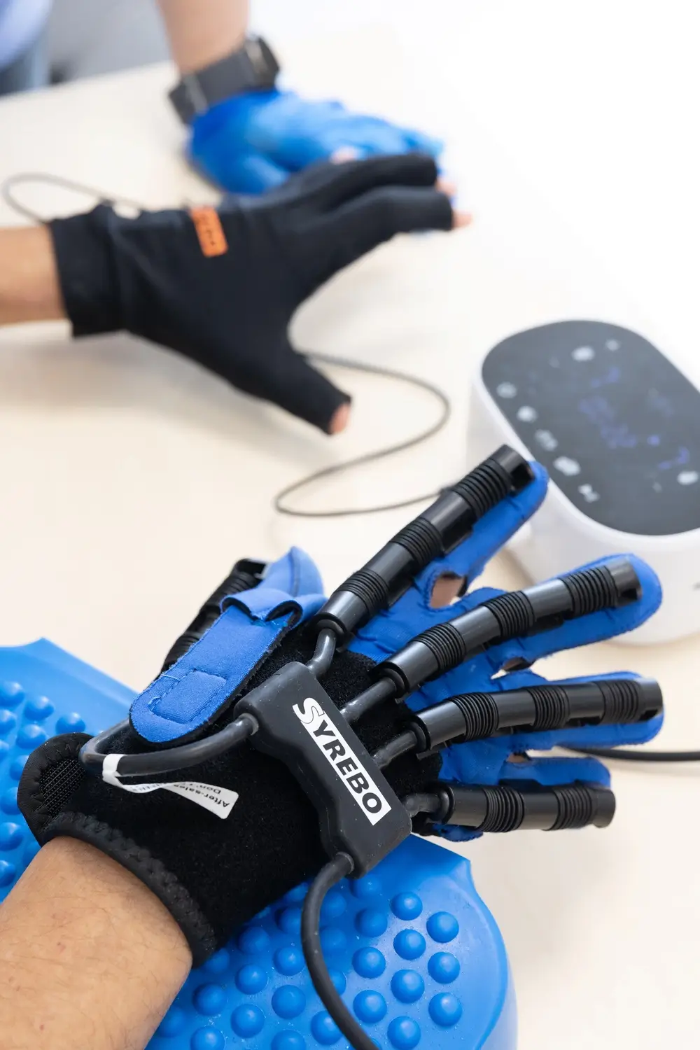 Close-up of both hands in robotic rehabilitation gloves resting on a textured blue ball beside a control unit