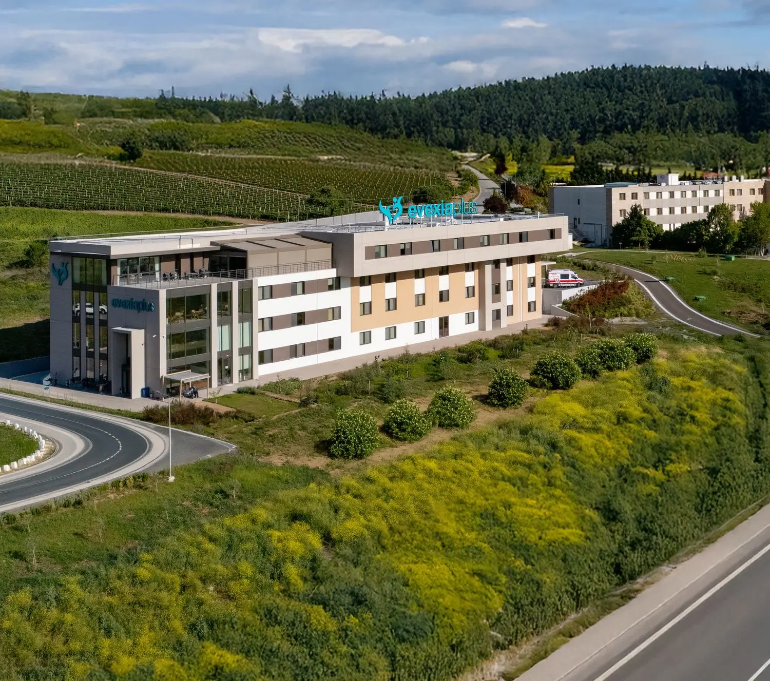 Aerial view of the Evexia Plus rehabilitation centre beside a curving road with hills and vineyards behind