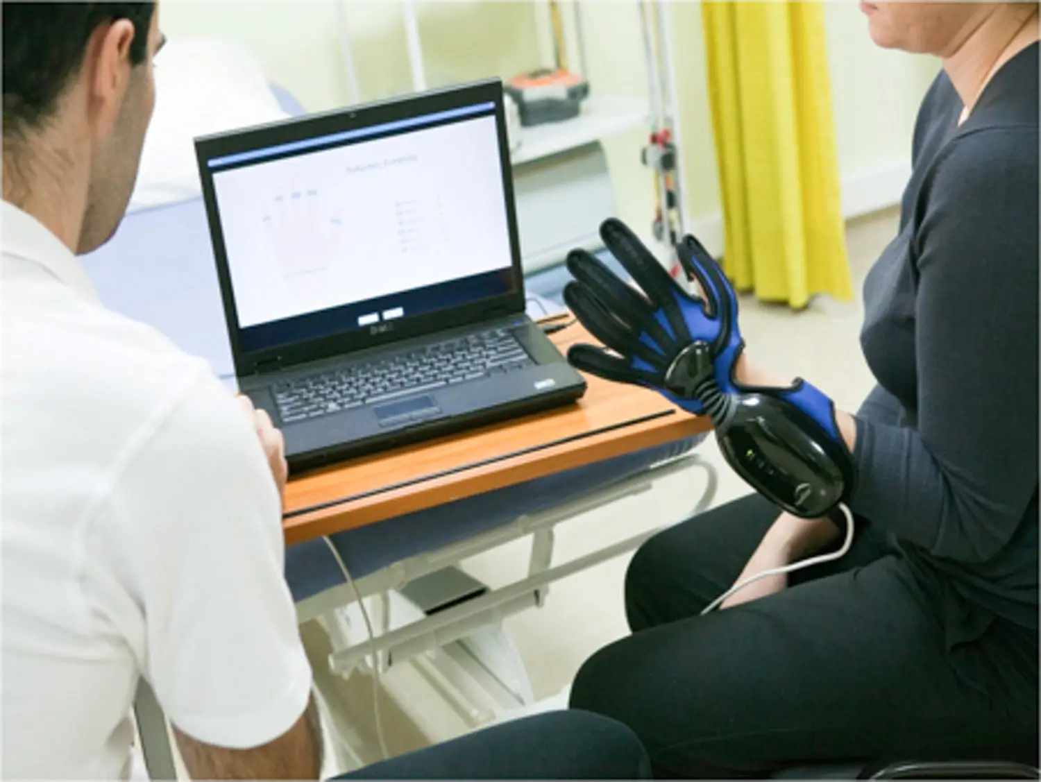 Clinician reviewing assessment data on a laptop while a patient wears a robotic hand glove