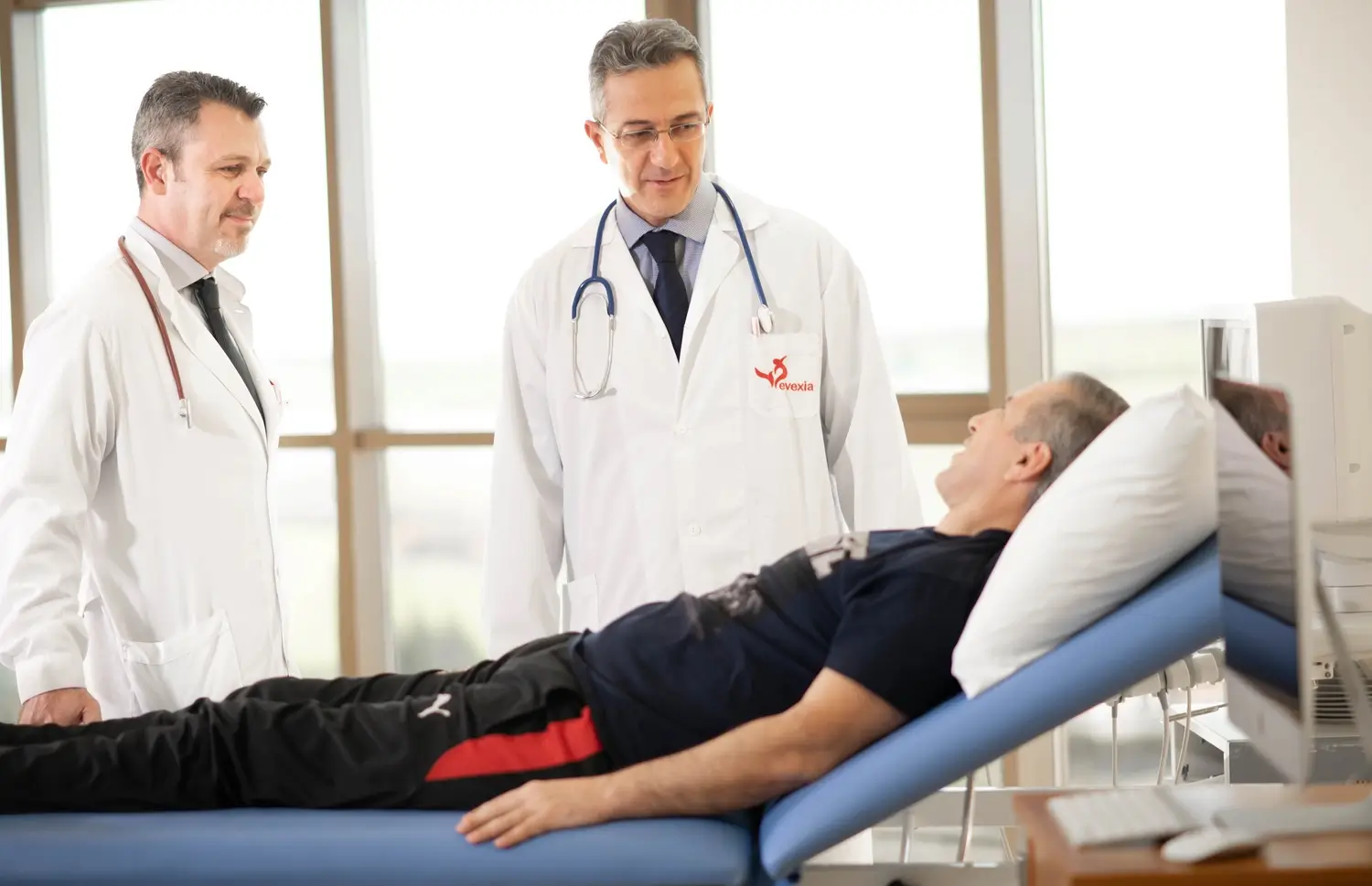 Two doctors in white coats talking with a man lying on a treatment couch beside a window