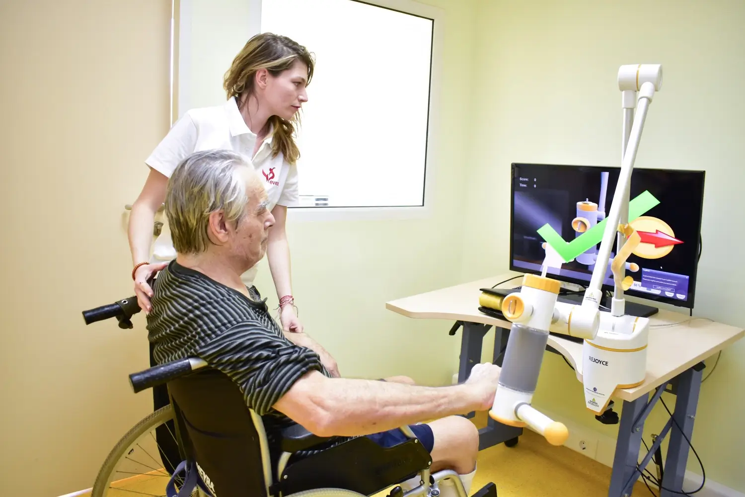 Therapist standing beside a man in a wheelchair using a robotic arm trainer in front of a screen