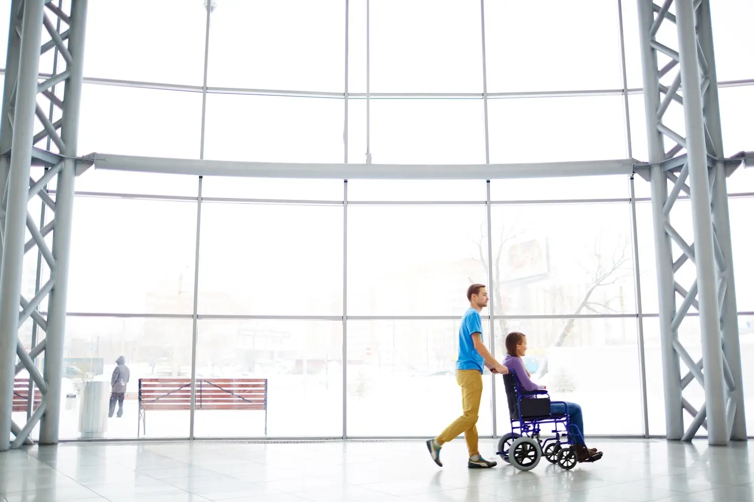 Staff member pushing a patient in a wheelchair across a bright atrium with a full-height glass wall