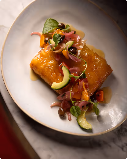 Golden brown square pastry topped with pickled onions, cucumber, greens, and a glossy sauce on a white plate.