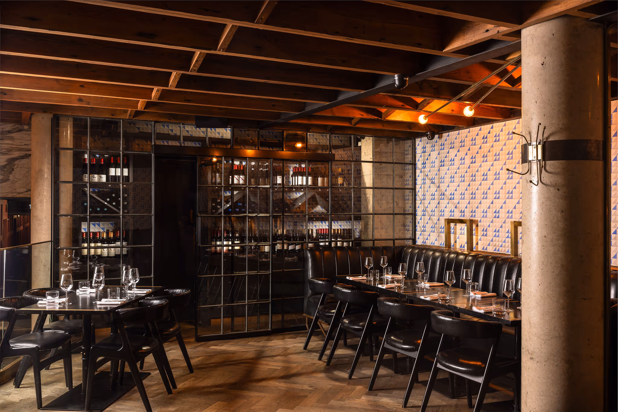 Elegant restaurant dining area with black leather chairs, wine storage, wood ceiling beams, and tiled wall.