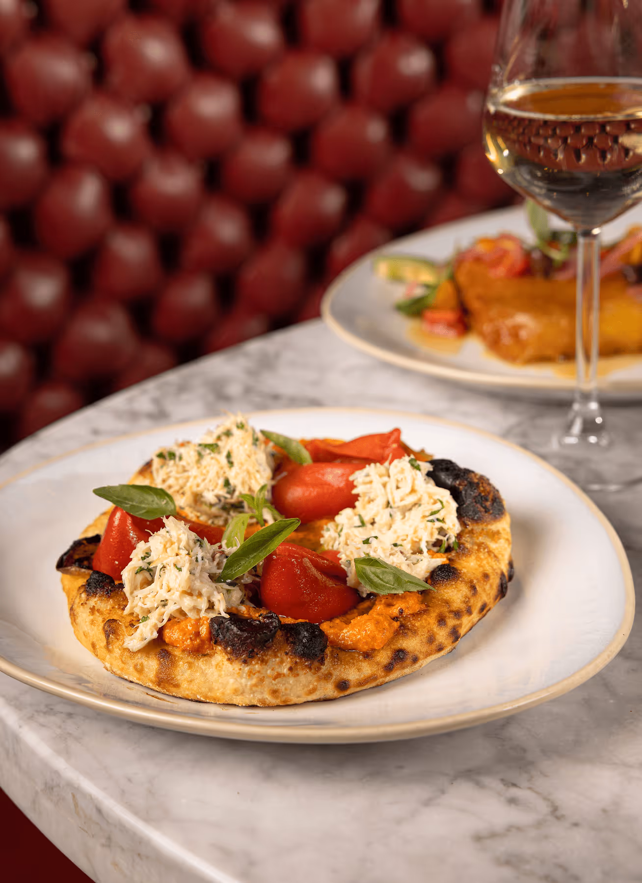 Charred flatbread topped with creamy crab, roasted red peppers, and basil leaves on a white plate.