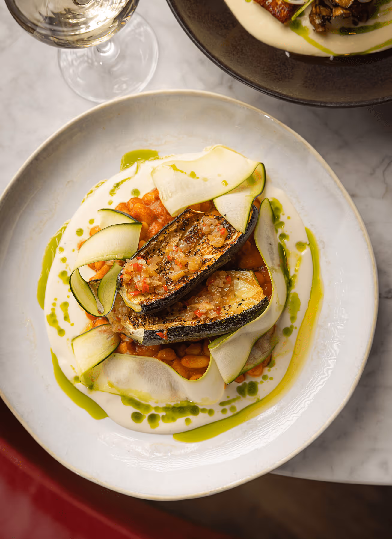 Grilled zucchini with beans, creamy sauce, and green herb oil on a white plate.
