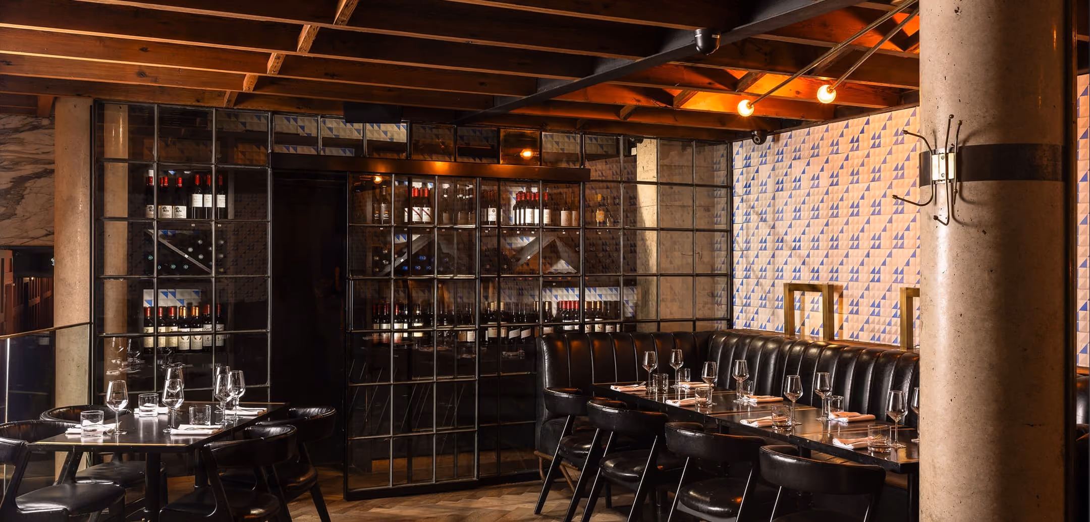 Cozy restaurant interior with black leather chairs, tables set with glasses, and a wine display behind glass panels.