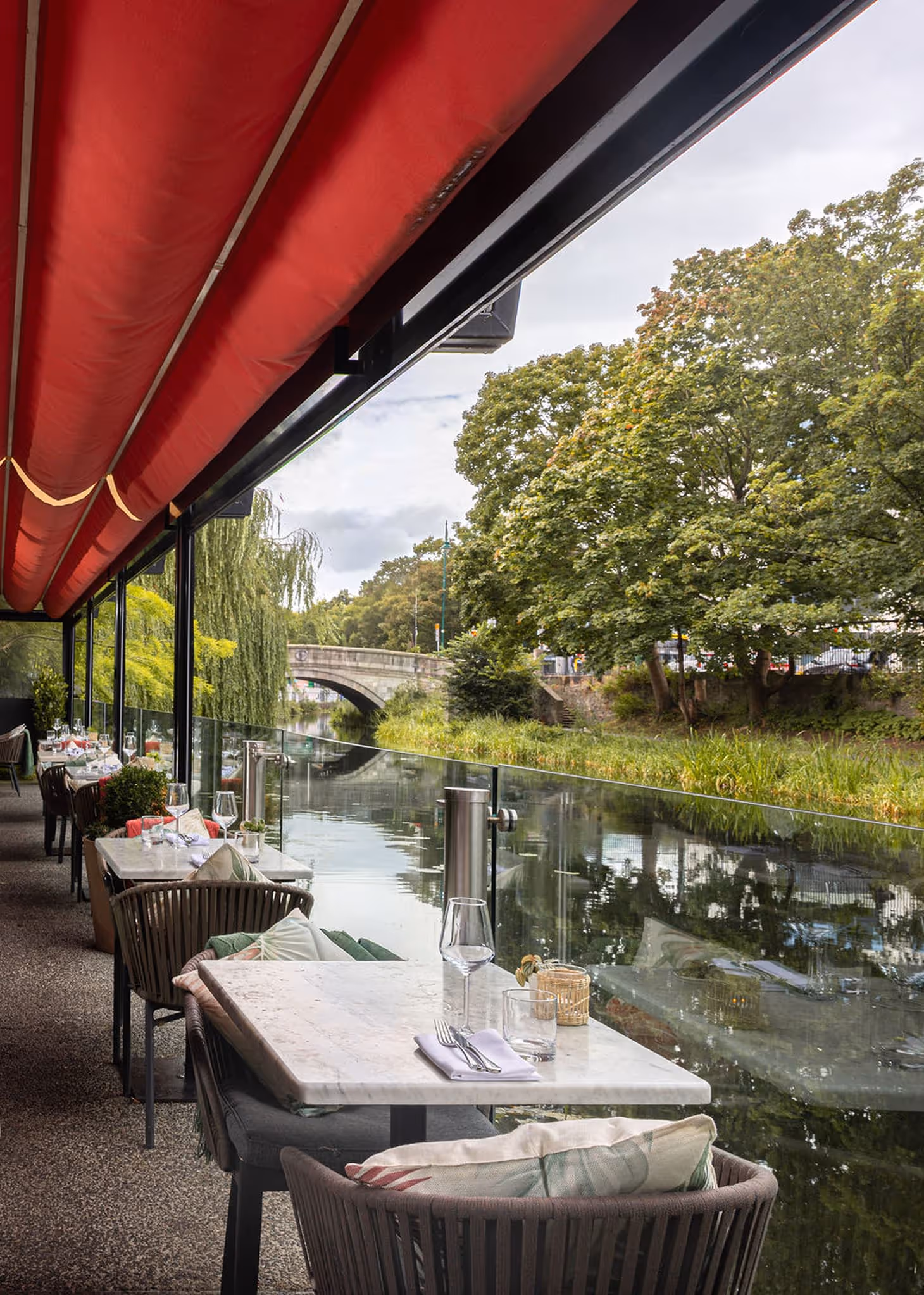 Outdoor canal-side dining area with tables, chairs, and red canopy overlooking trees and water.