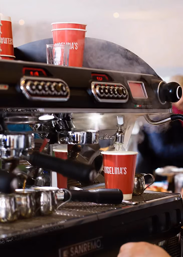 Espresso machine pouring coffee into a red Angelina's cup with steam in a cafe setting.