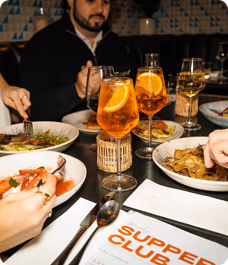 People dining with orange cocktails and plates of various foods on a dark restaurant table.