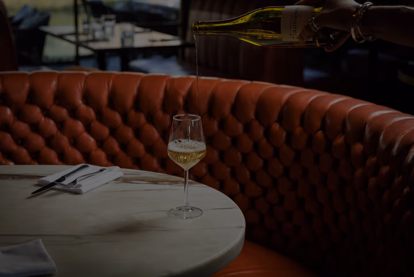 Wine being poured into a glass on a marble table with tufted brown leather seating in background.