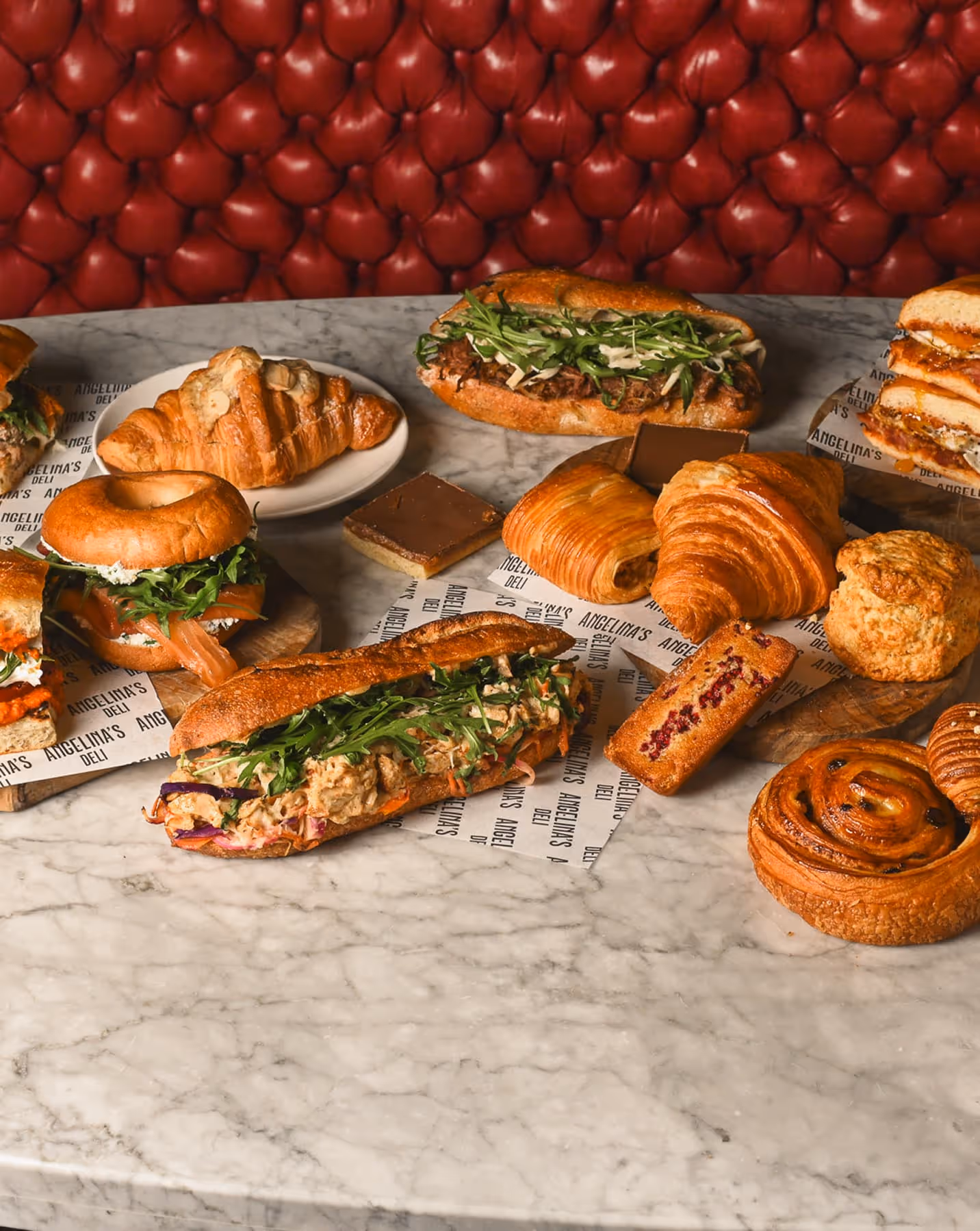 Variety of sandwiches, croissants, pastries, and baked goods on a marble table with red cushioned background.