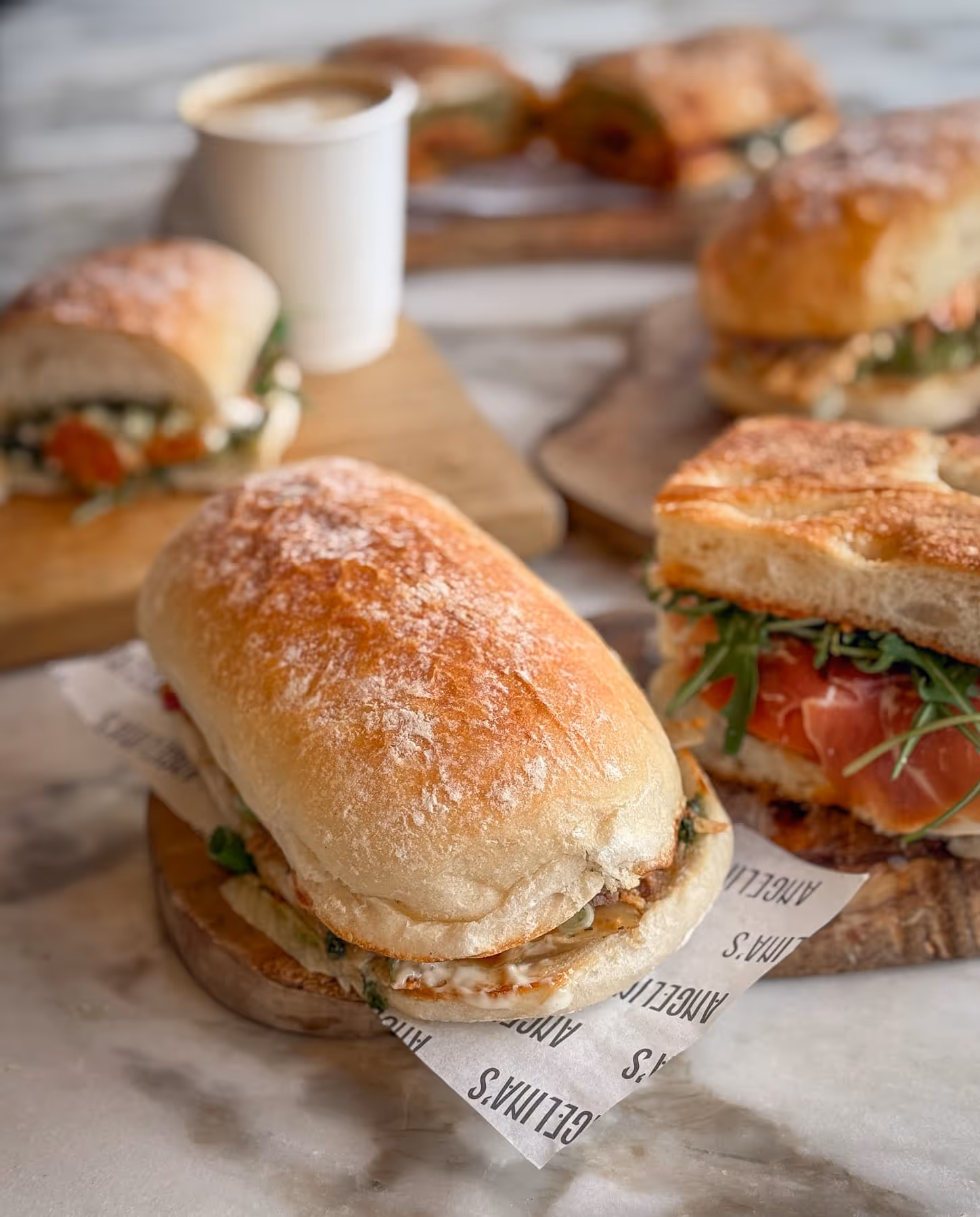 Various deli sandwiches on wooden boards with a cup of coffee in the background.