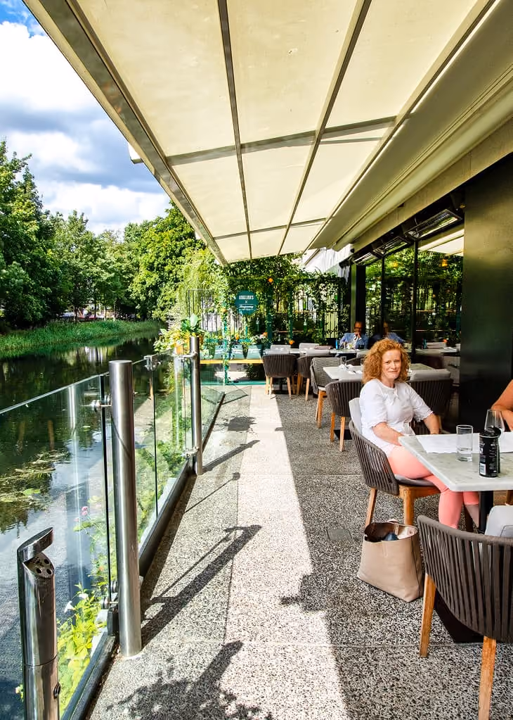 Outdoor dining terrace with tables, chairs, and glass railing overlooking a calm river and stone bridge.