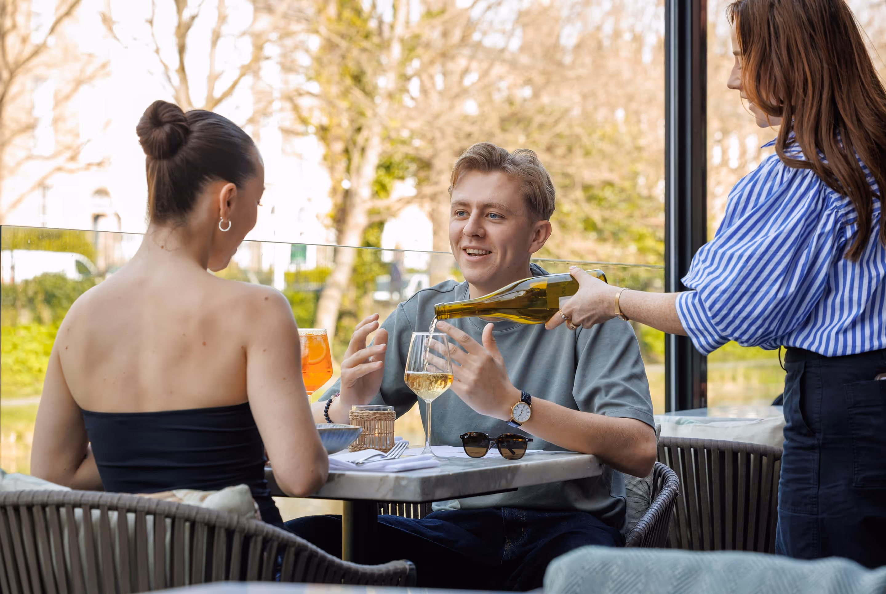 Woman pouring wine into a glass for a smiling man seated at an outdoor cafe table with another woman.