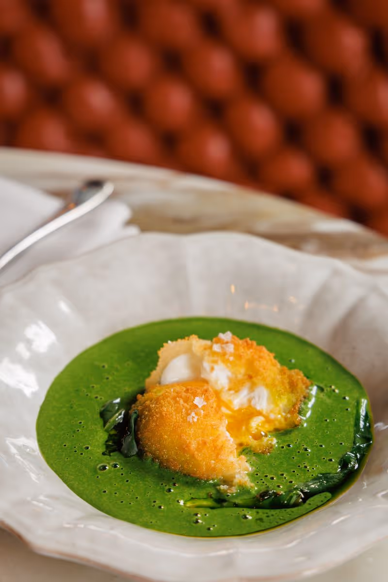 Breaded fried egg with runny yolk in vibrant green soup garnished with wilted greens on white plate.