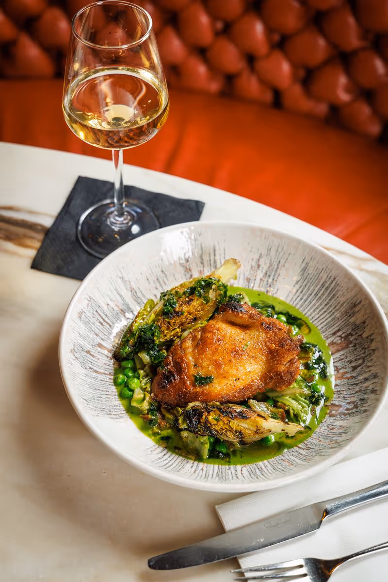 Plate with cooked chicken breast, grilled greens, peas in green sauce, and a glass of white wine.