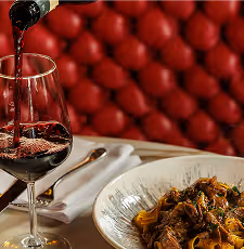 Red wine being poured into a glass next to a plate of pasta on a white tablecloth.