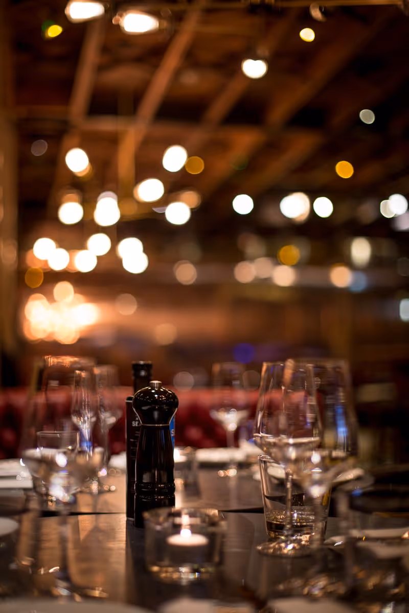 Close-up of a dining table with glasses, a pepper grinder, bottles, and bokeh lights in the background.