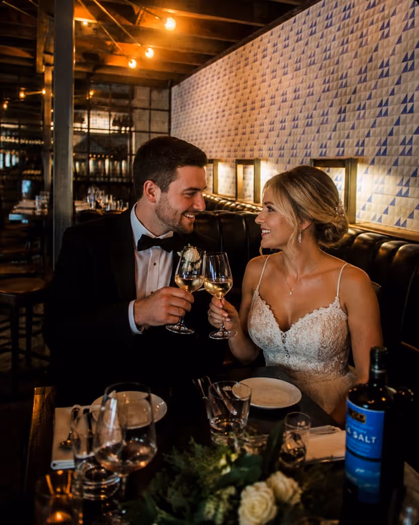 Bride and groom in wedding attire clinking wine glasses at a cozy restaurant table.