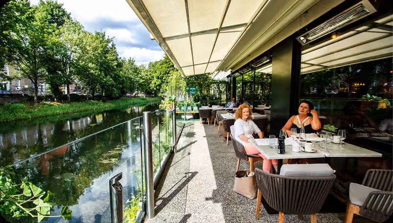 Two women sitting at a table on a riverside outdoor restaurant patio with trees and water view.