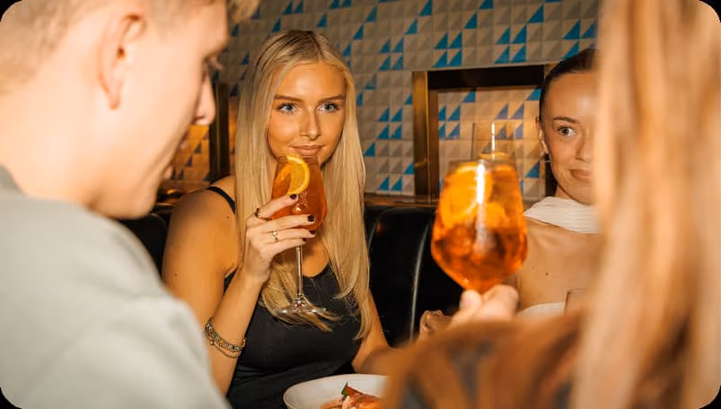 Three people sitting at a table, toasting with orange-colored cocktails with orange slices.