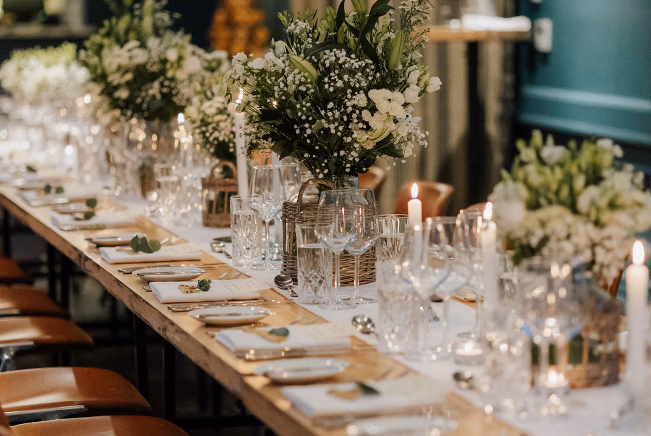 Long dining table with white floral centerpieces, lit candles, glassware, and place settings on wooden chairs.