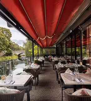 Outdoor restaurant seating along a river with tables set under a red canopy.