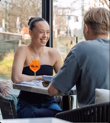 Smiling woman with a drink talks to a man at a table in a bright café.