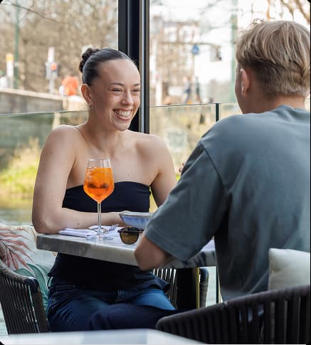 Smiling woman with a drink talks to a man at a table in a bright café.