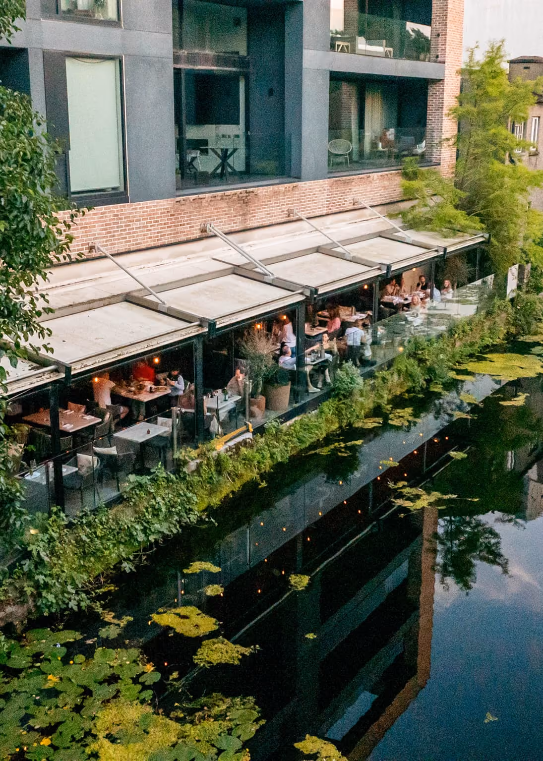 Outdoor restaurant seating along a river with plants and water lilies reflecting nearby buildings.