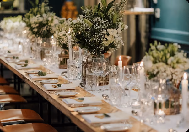 Long wooden table set with white napkins, glassware, candles, and green floral centerpieces for a celebration.