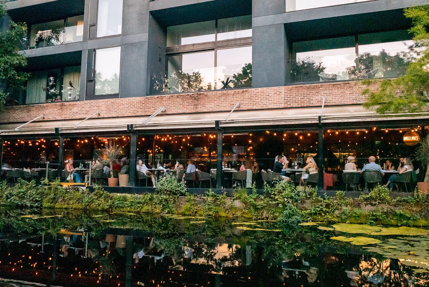 People dining on a lit outdoor patio of a restaurant beside a pond with green plants.