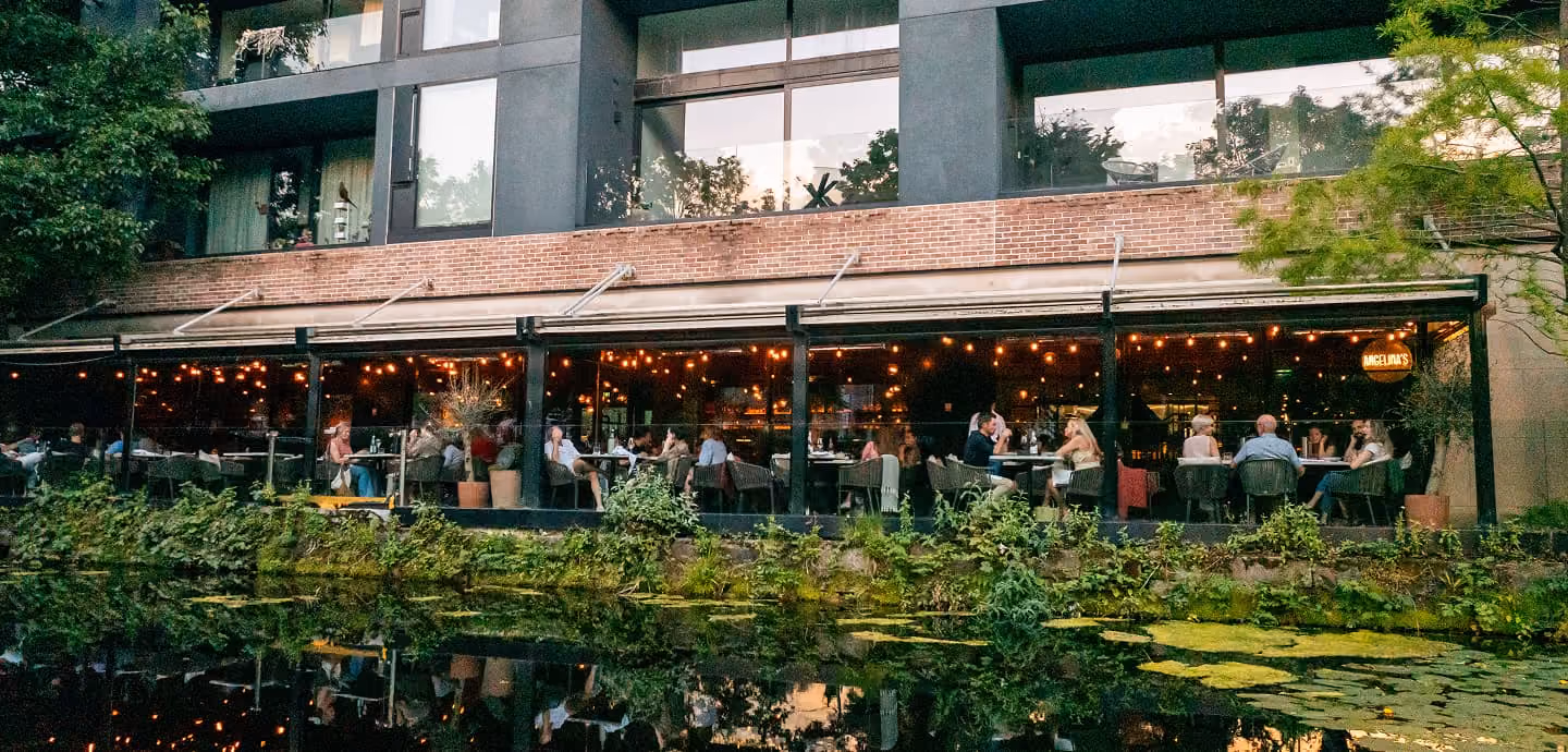 Outdoor dining area with people seated at tables under string lights beside a pond with greenery.