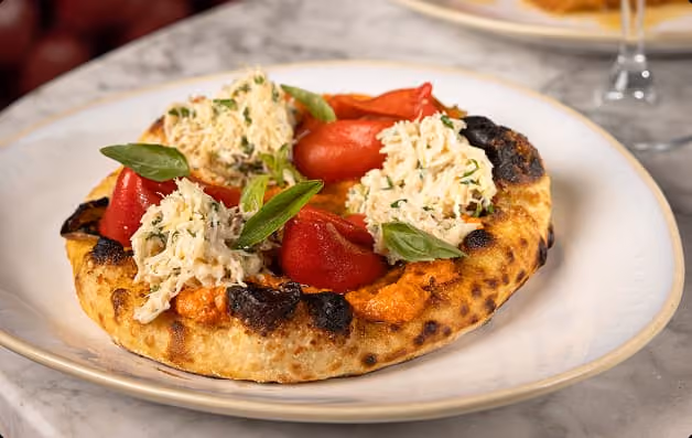 Small pizza with tomato sauce, red roasted peppers, crab meat, and fresh basil on a white plate.