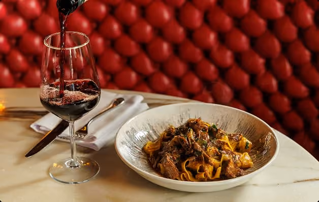 Glass of red wine being poured next to a plate of pasta with beef on a marble table.