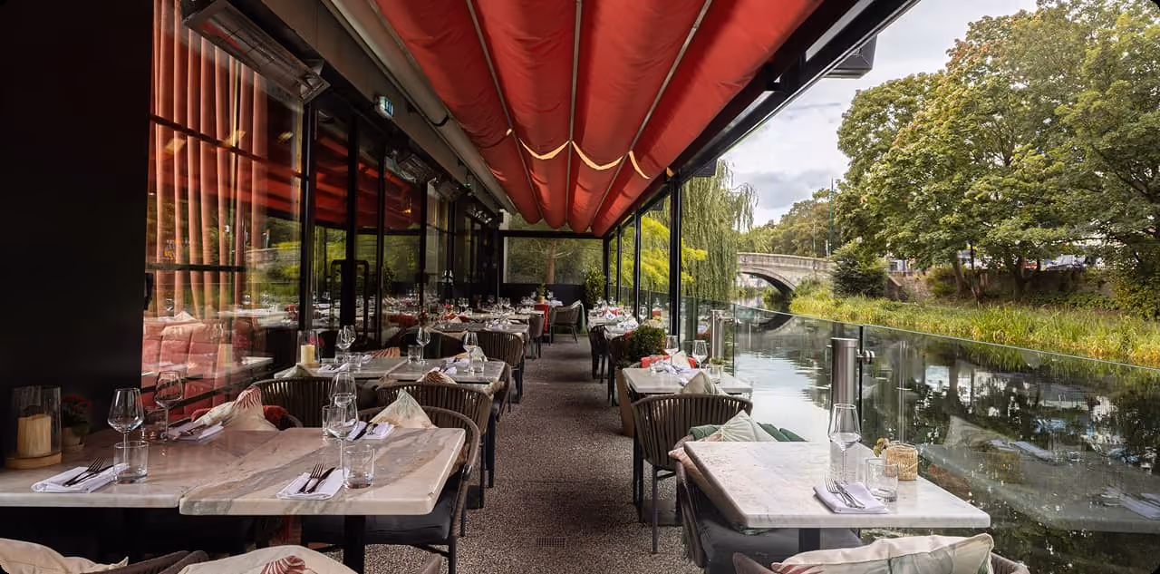 Outdoor restaurant seating beside a river with tables set under a red canopy.