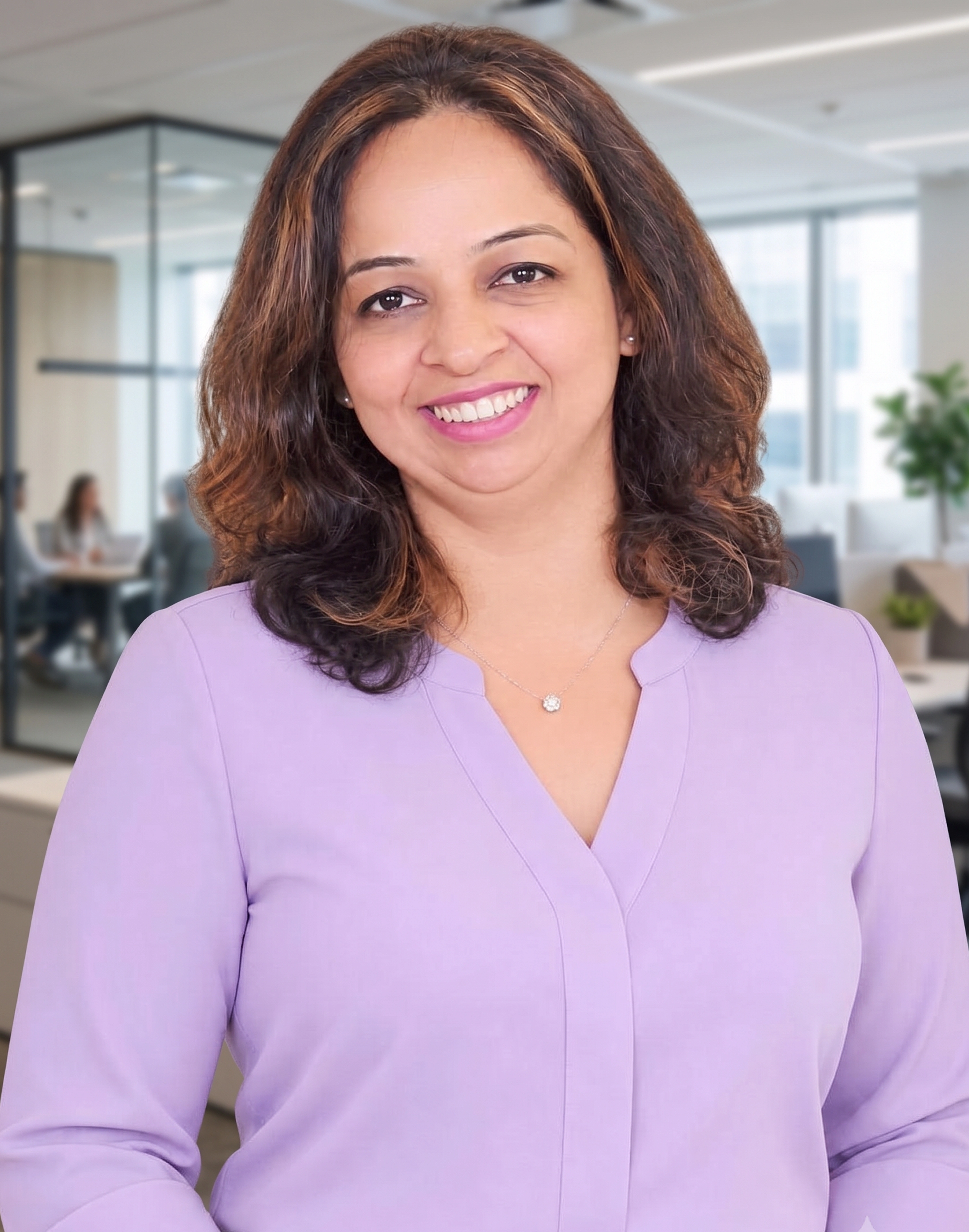 Smiling woman with shoulder-length wavy brown hair wearing a light purple blouse and a small pendant necklace in a modern office setting with blurred coworkers and windows in the background.