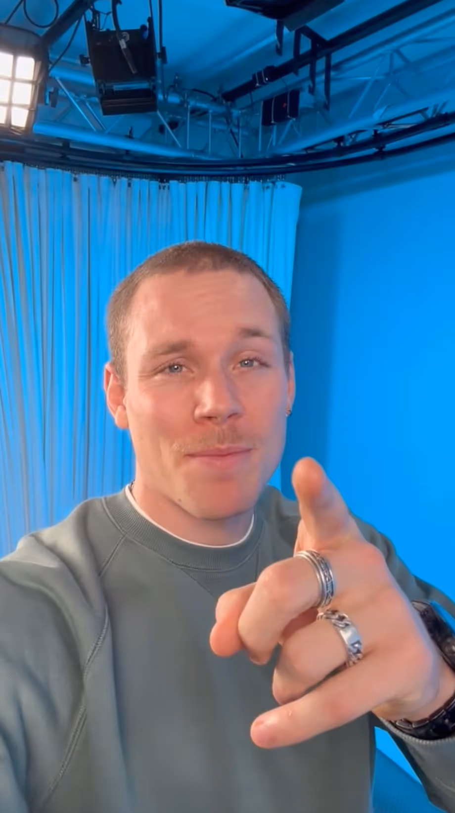 Man in green sweatshirt points directly at the camera with a slight smile, blue background behind him.