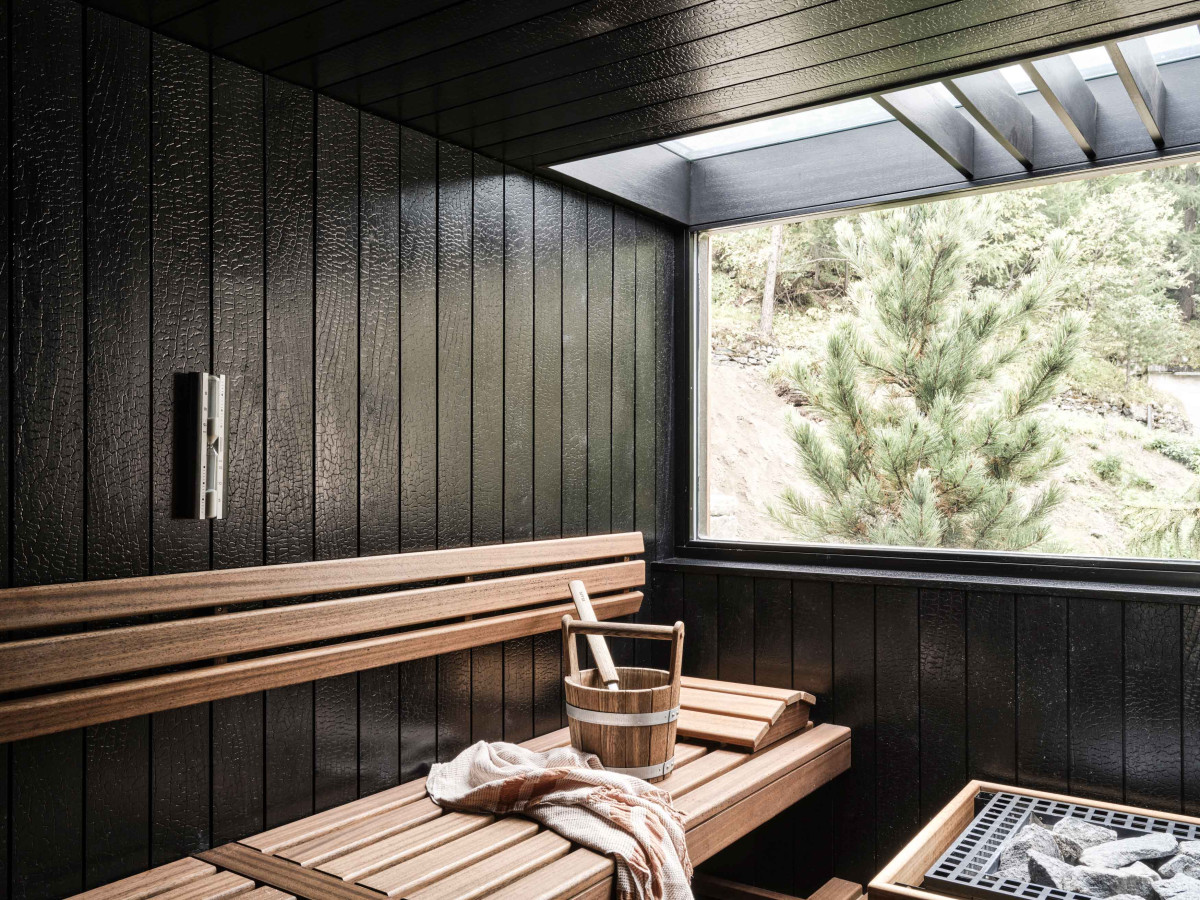 Intérieur épuré du sauna en bois sombre de l'Atman Mountain Spa au CERVO Mountain Resort, avec un seau traditionnel et une large ouverture vitrée sur les sapins de Zermatt.