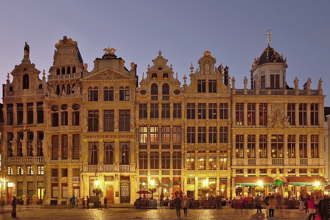 Façades historiques de la Grand-Place de Bruxelles à l’heure bleue, avec quelques passants. Photo Benoit Brummer, CC BY 4.0, recadrée et légèrement désaturée.