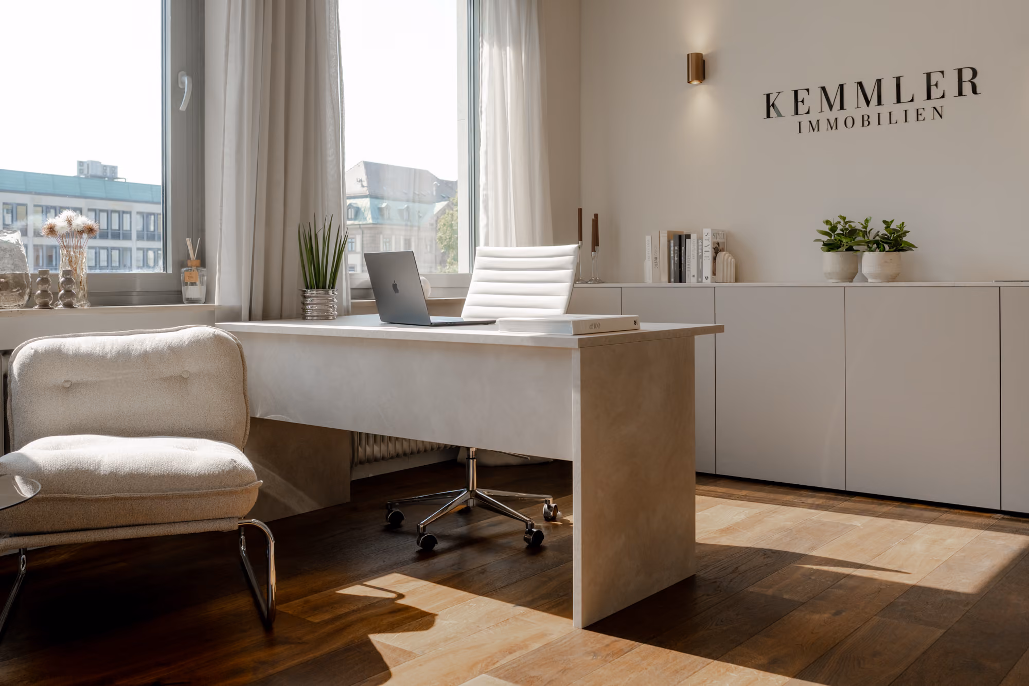 Bright modern office with white desk, chair, laptop, armchair, plants, and Kemmler Immobilien sign on wall.
