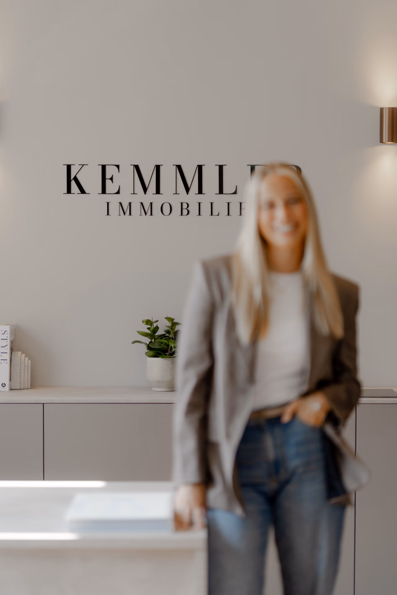 Blurred woman in casual businesswear stands inside an office with 'Kemmler Immobilien' sign.
