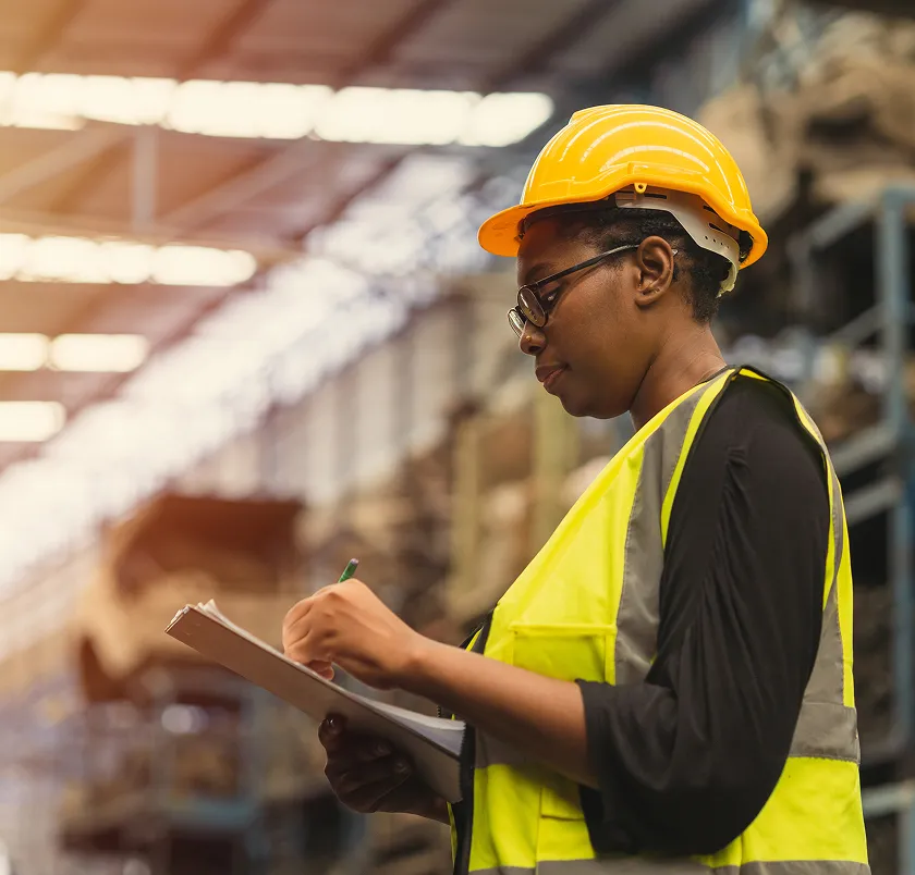 Worker in a hard hat writing on a clipboard in a warehouse